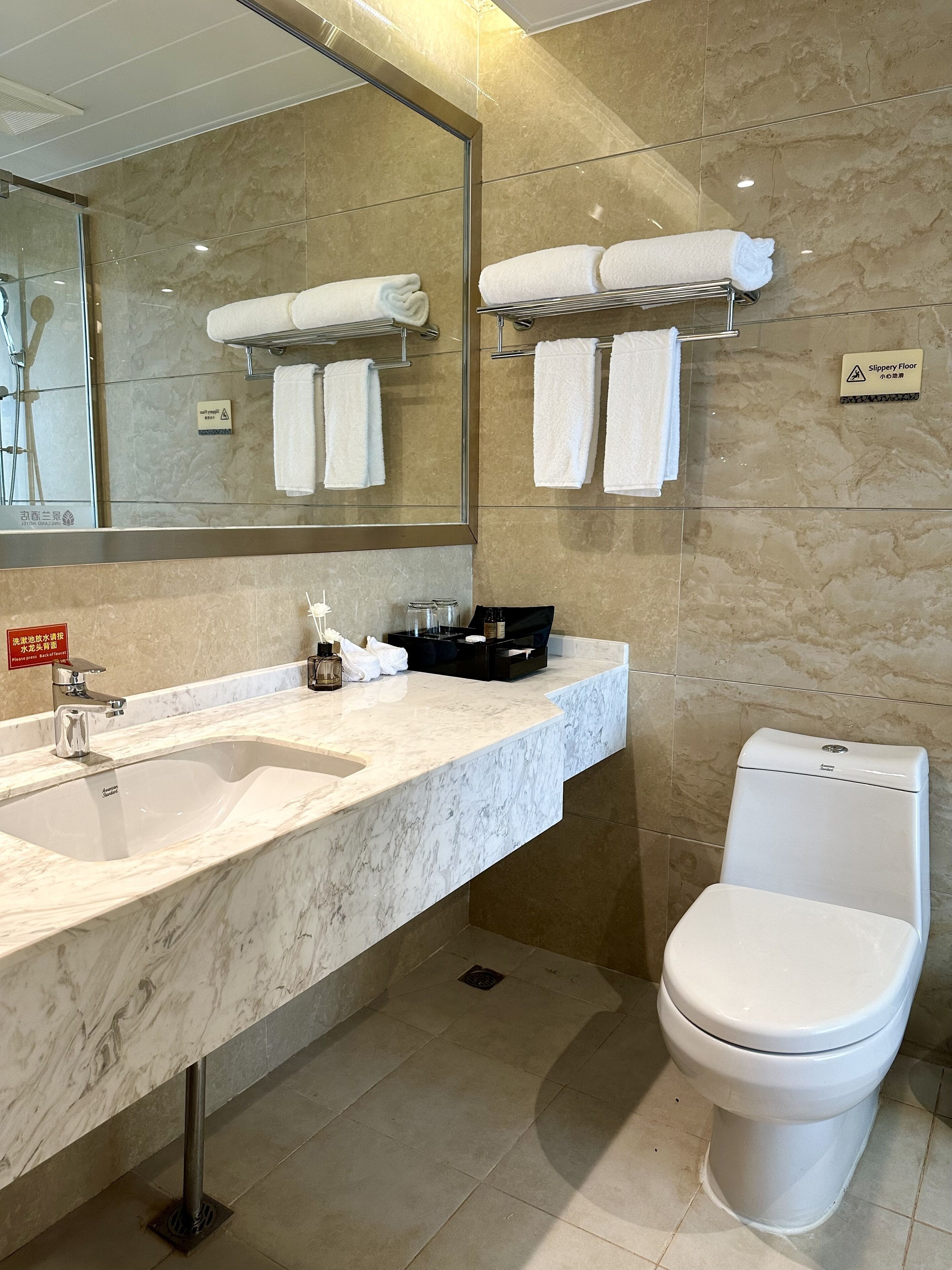 A modern bathroom with a marble sink, a toilet, and a mirror. There are towels hanging on a rack and a sign indicating a slippery floor.