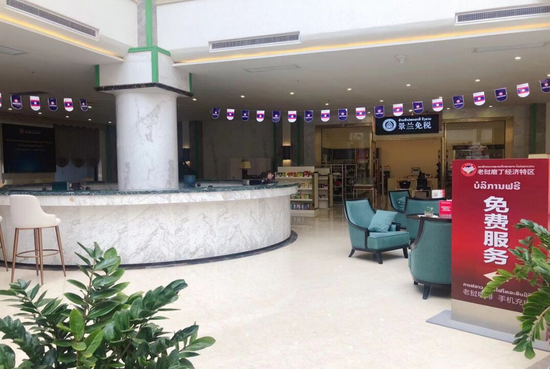 A lobby with a reception desk, seating area, and a large pillar. There are flags hanging from the ceiling and a sign with text in a foreign language.
