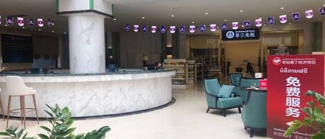 A lobby with a reception desk, seating area, and a large pillar. There are flags hanging from the ceiling and a sign with text in a foreign language.
