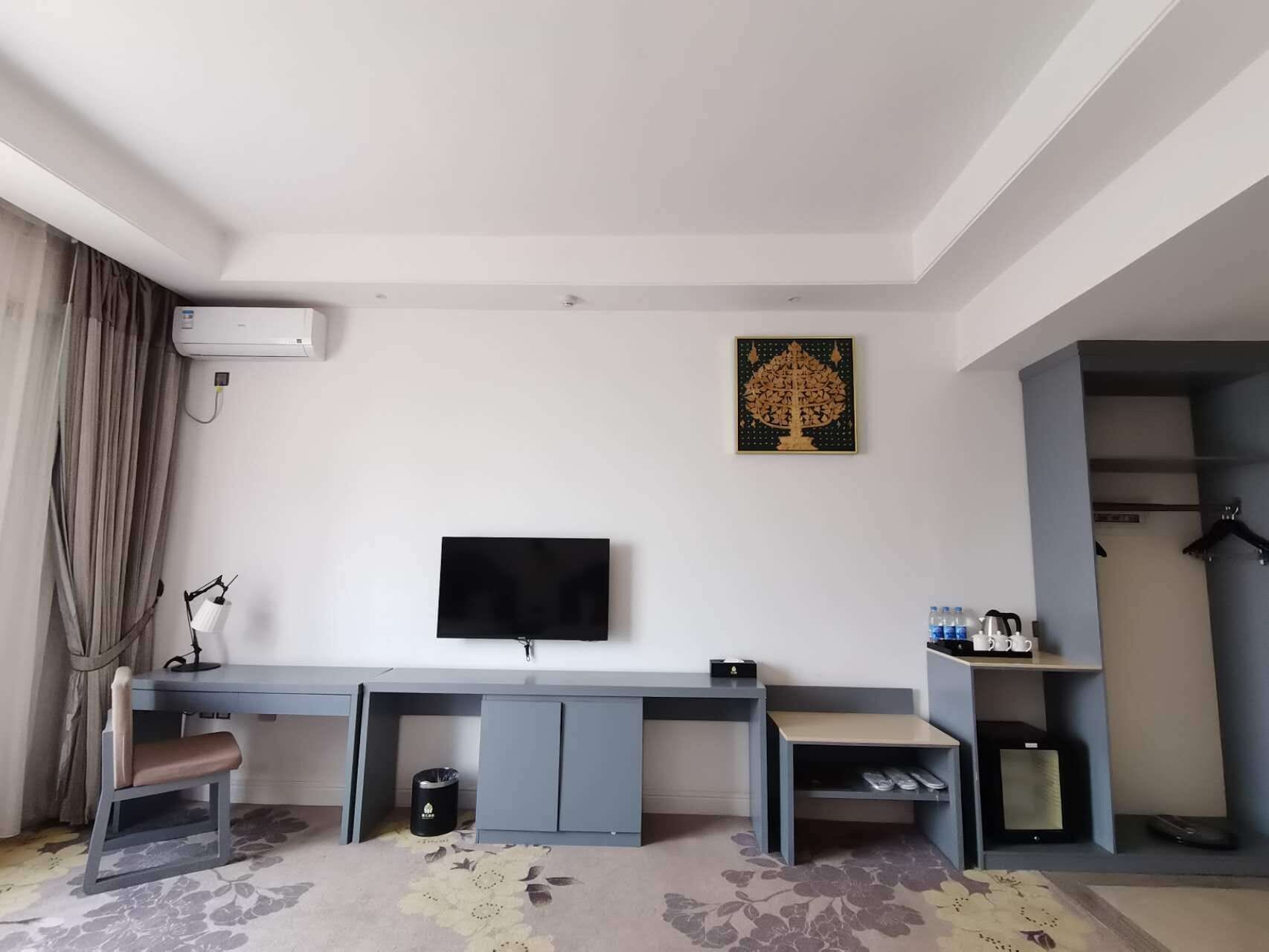 A hotel room with a flat-screen TV, a desk with a chair, a small trash bin, and a shoe rack.