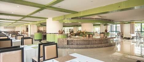 A spacious hotel lobby with multiple dining tables and chairs, a reception area, and a central fountain.
