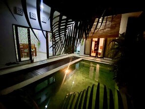 A swimming pool with a wooden deck, a glass door leading to a room, and a large plant in the background. - Villa Sunny Melati Canggu (Canggu)