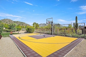 Quadra de basquete ao ar livre com rede, cercada por uma grade e cactos.