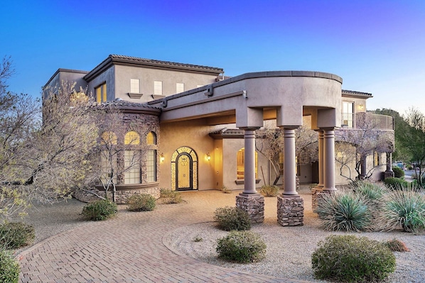 A large, modern house with a curved entrance, stone pillars, and a well-maintained garden with various plants.