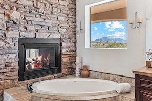 A bathroom with a stone wall, a fireplace, a large bathtub, and a window with a mountain view.
