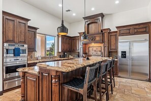 Una cocina con gabinetes de madera, una isla grande, taburetes de bar, un refrigerador de acero inoxidable e iluminación colgante.