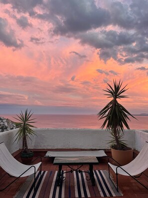 Une terrasse avec vue sur la mer, un banc, deux plantes en pot et une rambarde blanche.