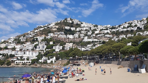 Une plage où des gens se prélassent au soleil et se baignent, un rivage sablonneux et, au loin, sur une colline, des bâtiments blancs.