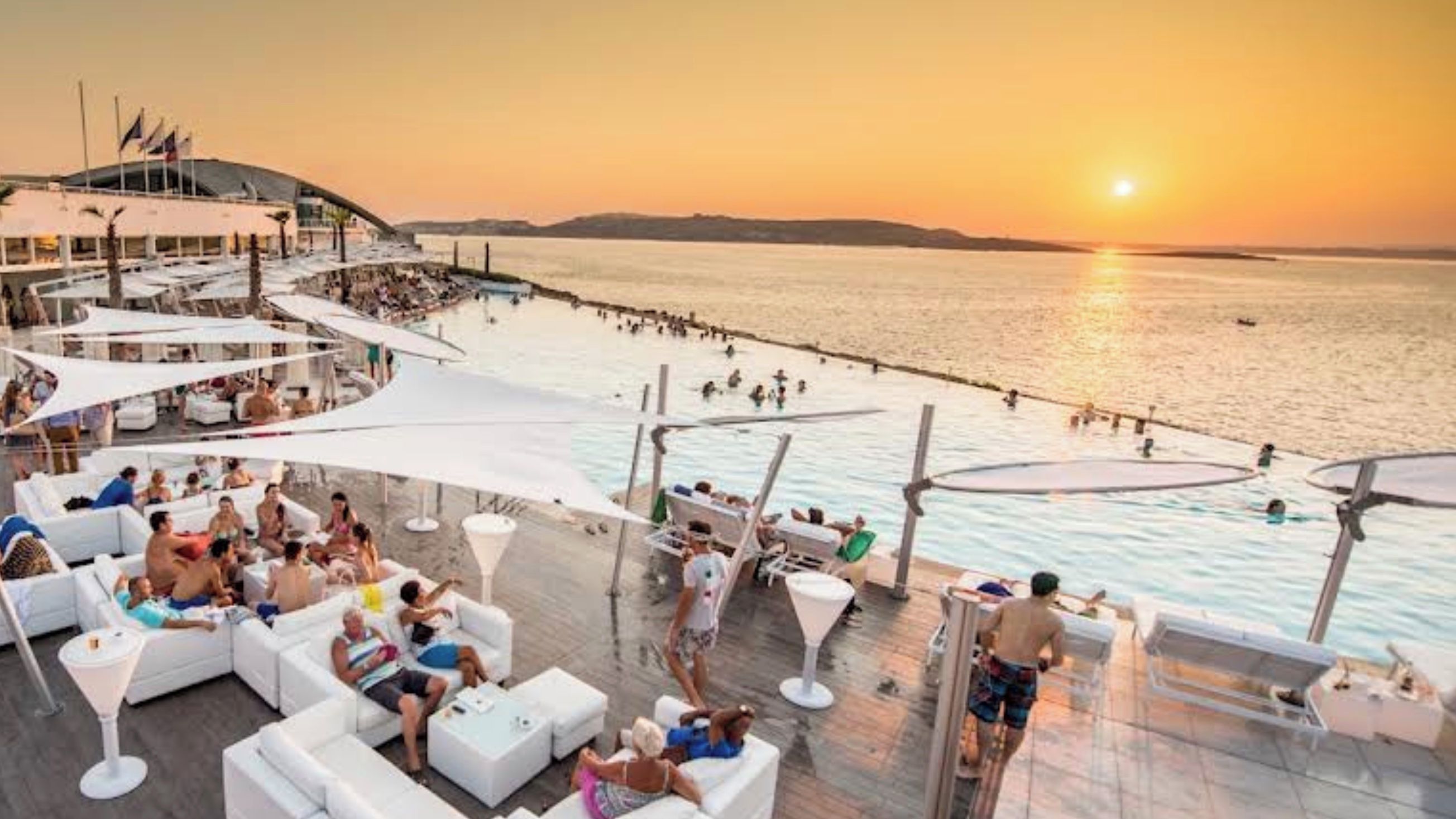 A rooftop bar with outdoor seating and a view of the sea at sunset.
