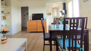 A modern living room with a wooden dining table, chairs, a TV, and a kitchen in the background.