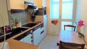 A compact kitchen with white cabinets, a microwave, and a sink. There is a dining table with chairs and a potted plant.