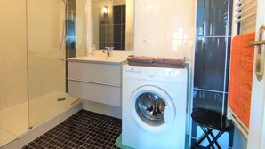 A compact bathroom with a shower, a sink, a mirror, a washing machine, and a towel rack.