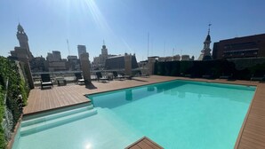 Una piscina en la azotea con terraza de madera y sillas reclinables, rodeada de edificios de la ciudad y un cielo despejado.