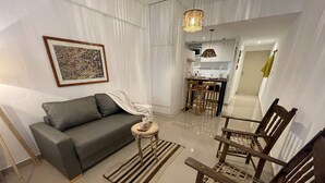 Una sala de estar con sofá, mesa de centro y una silla. Se ve una cocina al fondo.