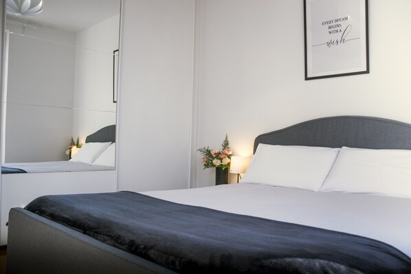 A modern hotel room with a large bed, a bedside table with a lamp, a framed inspirational quote on the wall, and a mirror reflecting the room.