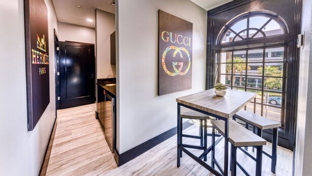 Ein modernes Interieur mit Holzboden, einem Tisch mit Stühlen, einem großen Fenster mit Blick auf Palmen und einer Wand mit Gucci-Logo.