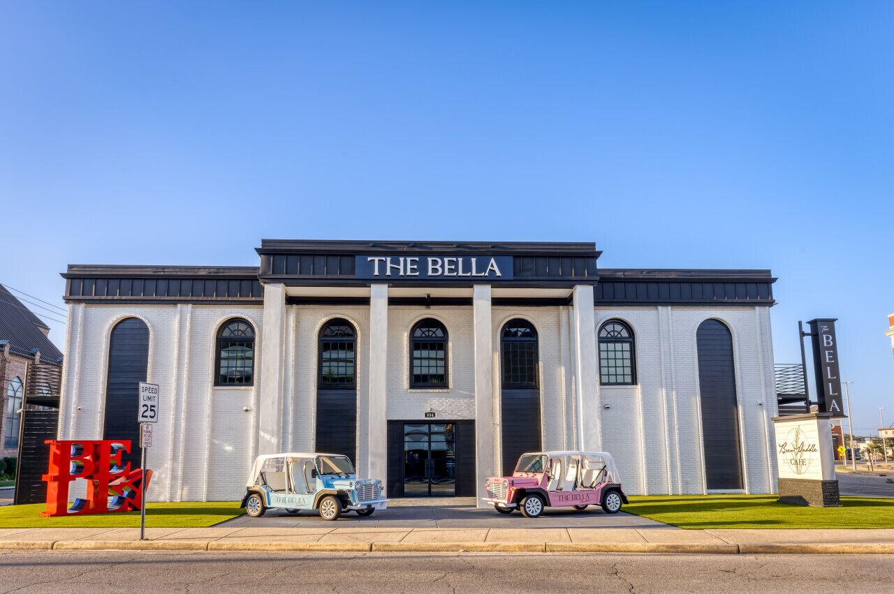 Un edificio con un cartel que dice "THE BELLA" y dos coches antiguos estacionados adelante.