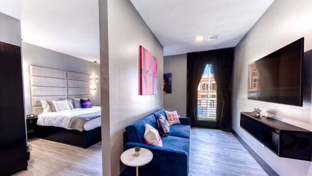 Quarto de hotel moderno com uma cama, um sofá azul, uma mesa redonda pequena e uma TV fixada na parede.