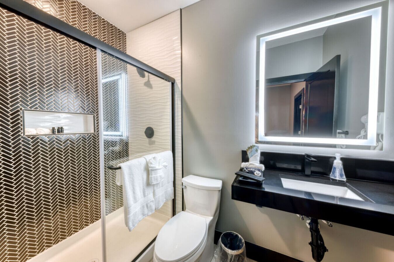 A modern bathroom with a herringbone-patterned wall, a glass shower enclosure, a white toilet, a black sink with a mirror, and a trash can.