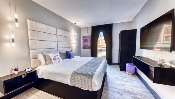 A modern bedroom with a large bed, a flat-screen TV, and a view of a building through a window. - The Bella`s Purple Prince Suite, Sleeps 2, Private Balcony (Biloxi)