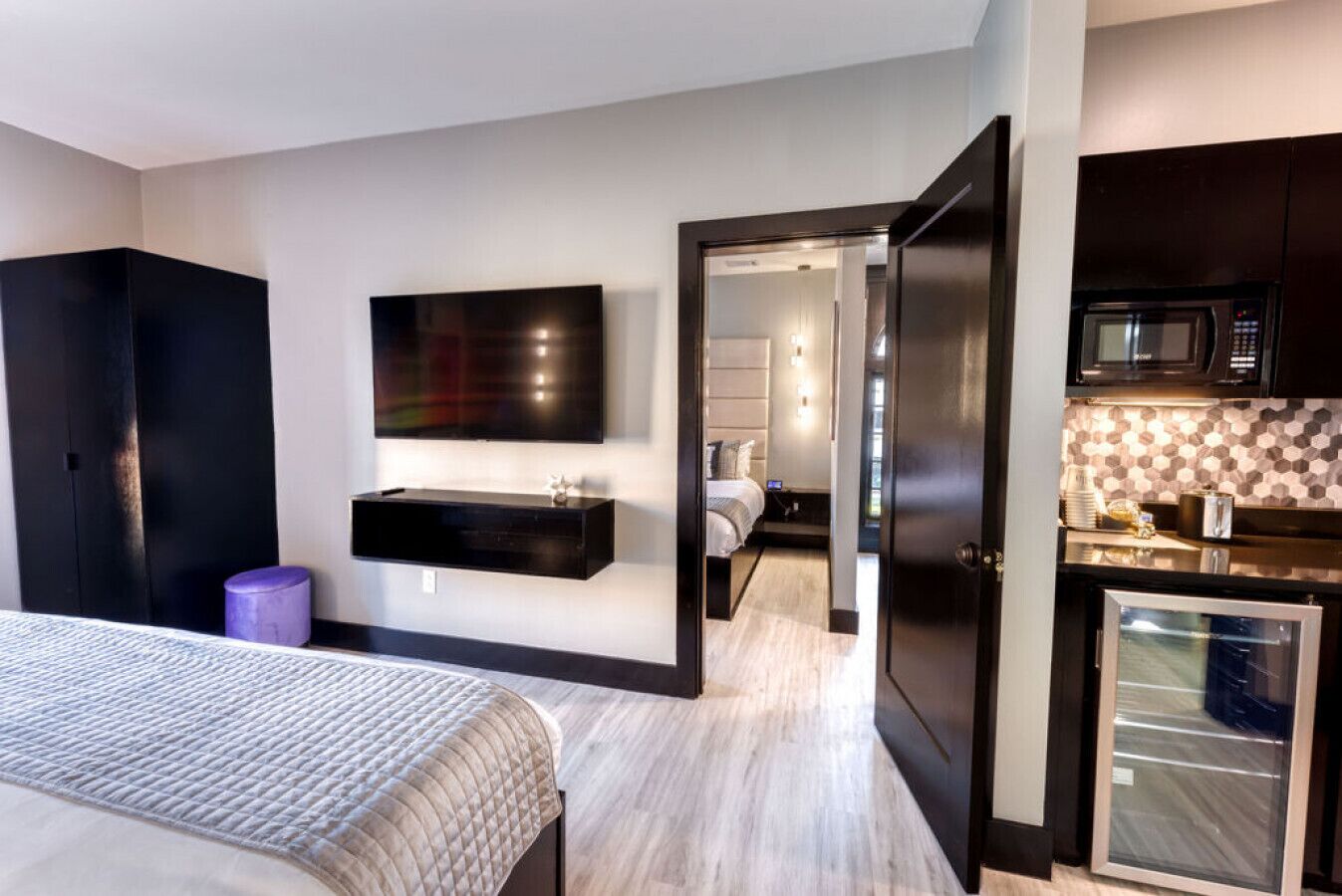 Ein modernes Hotelzimmer mit einem Bett, einem Flachbildfernseher, einer integrierten Sitzbank, einem kleinen violetten Hocker und einer Küchenzeile mit Mikrowelle und Kühlschrank.