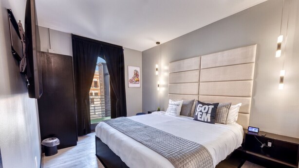Ein modernes Hotelzimmer mit einem großen Bett, einem flachbildfernseher an der Wand und einem kleinen Nachttisch mit Ladestation.