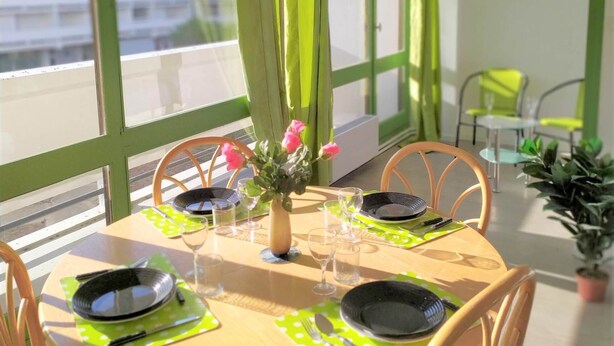 Uma mesa de jantar redonda com jogos americanos verdes, pratos pretos e copos de vidro transparente. Cadeiras de madeira circundam a mesa. Um vaso com flores cor-de-rosa está sobre a mesa.
