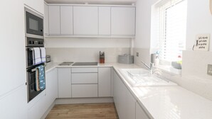 A modern kitchen with white cabinetry, a stainless steel oven, a microwave, and a window with blinds. - Llwyn Onn (Pwllheli)