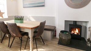 A dining area with a wooden table, two chairs, pendant lights, a potted plant, a painting, a mirror, and a fireplace with a log fire. - Llwyn Onn (Pwllheli)