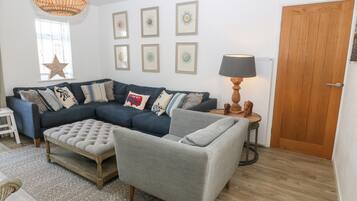 Sala de estar com um sofá azul escuro, uma poltrona cinza, uma mesa de centro de madeira e um abajur sobre uma mesinha lateral. Há quadros nas paredes e uma porta de madeira ao fundo.