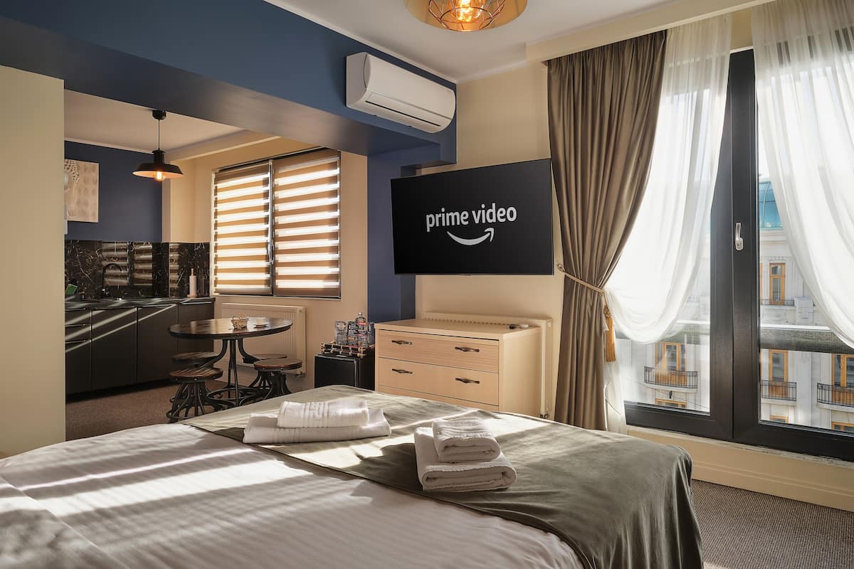 A hotel room with a bed, a TV displaying the Prime Video logo, a kitchen area, and a view of the city through large windows.