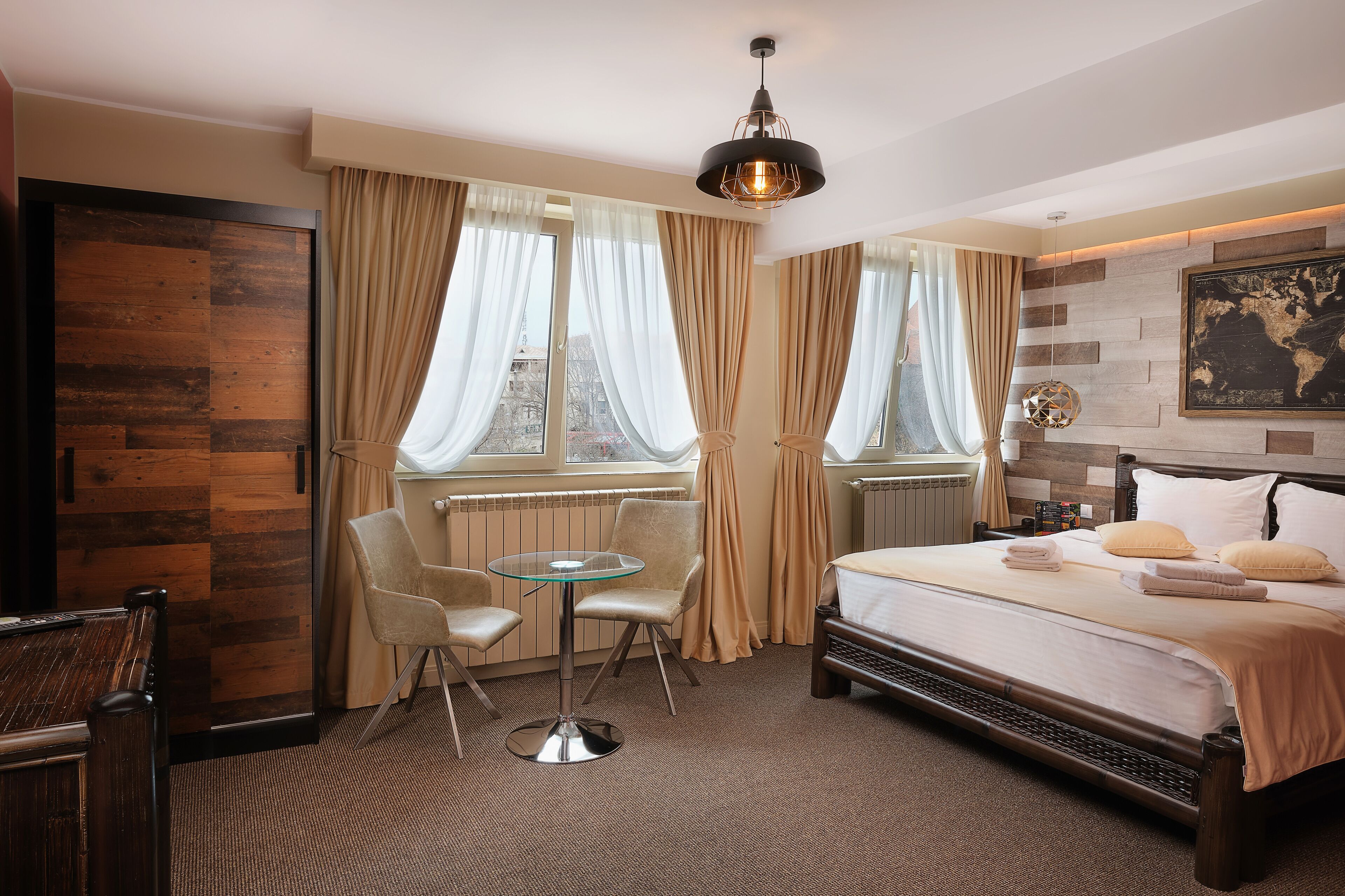 A hotel room with a bed, a glass table, two chairs, a view of the city through large windows, and a modern light fixture.