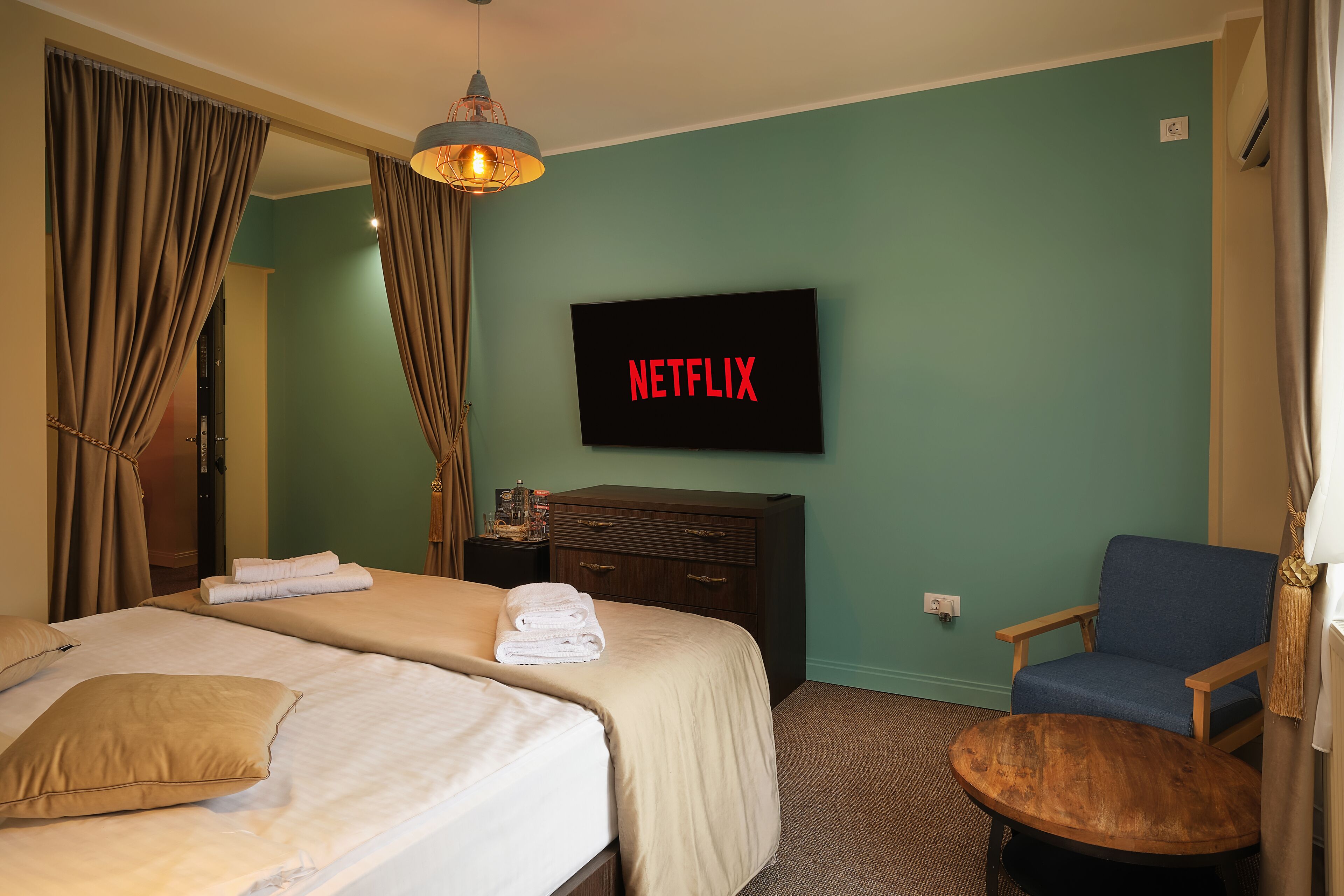 Ett hotellrum med en säng, en stol, en tv som visar ”NETFLIX” och ett litet bord i trä.
