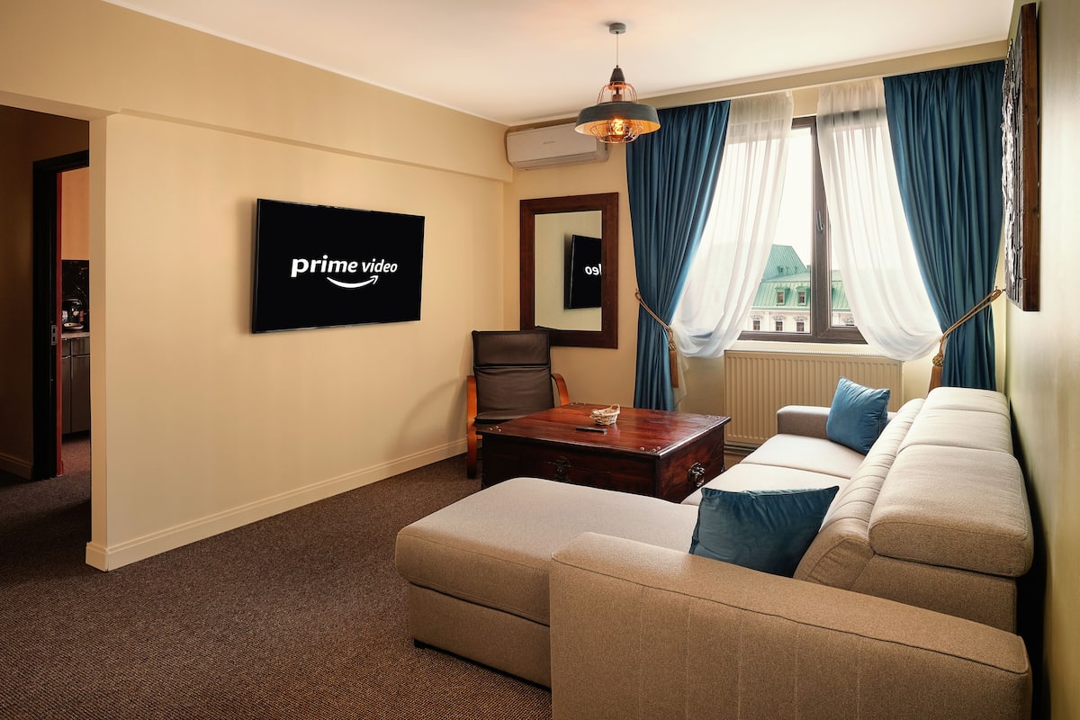 A living room with a flat-screen TV displaying 'prime video', a sofa, a coffee table, and a view of a building through a window with blue curtains.