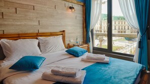 A hotel room with a wooden headboard, a large bed with white and blue bedding, a nightstand with a water bottle, and a view of buildings through the window. - Piata Unirii Central Apartments (Iasi)