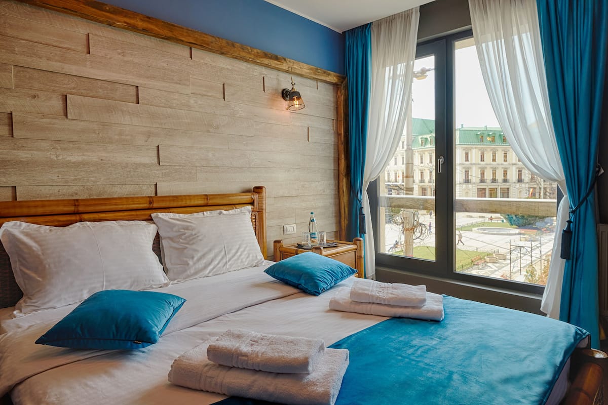 A hotel room with a wooden headboard, a large bed with white and blue bedding, a nightstand with a water bottle, and a view of buildings through the window.