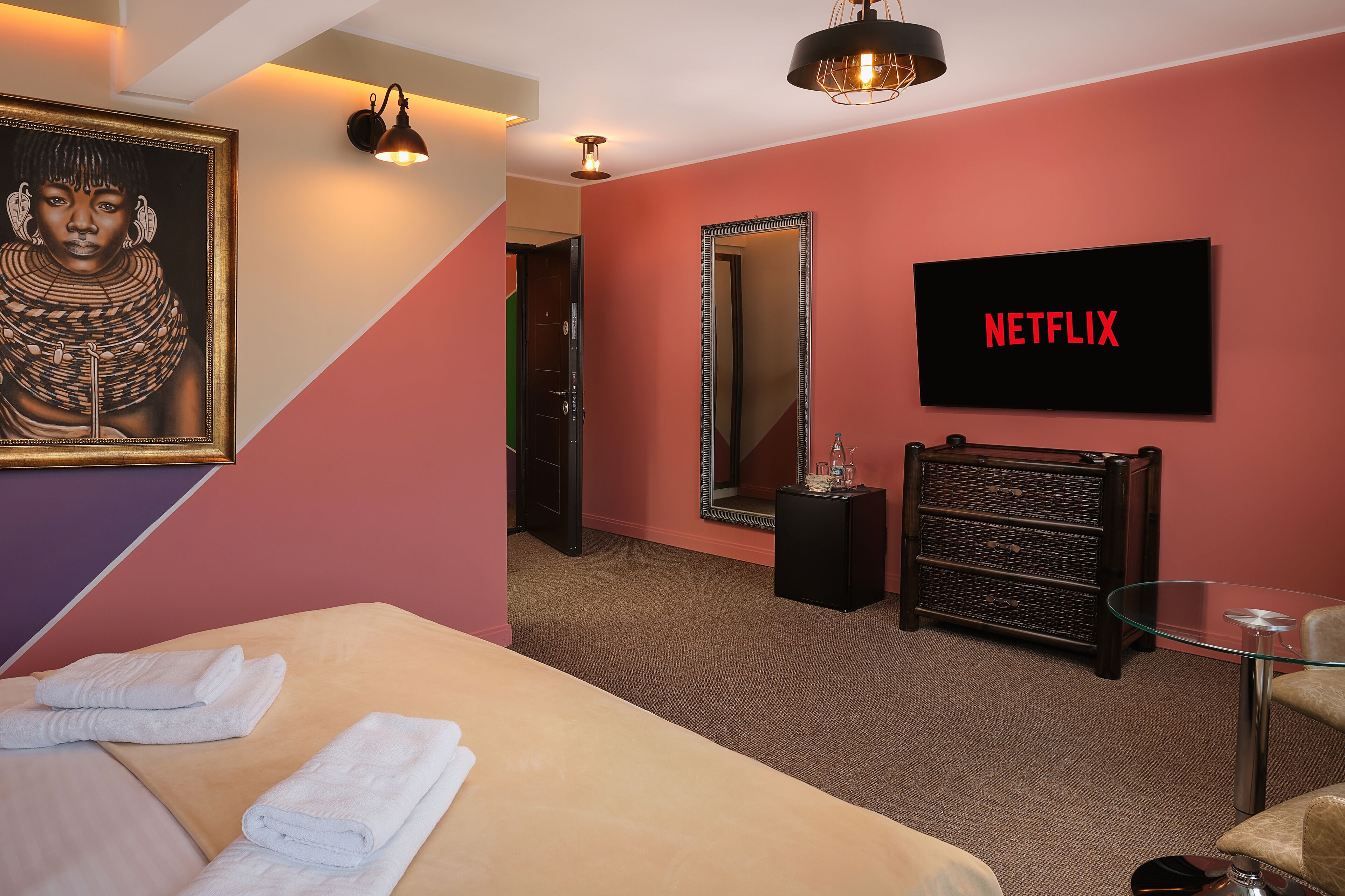 A hotel room with a bed, a glass table, a mirror, a television displaying 'NETFLIX', and a framed artwork on the wall.