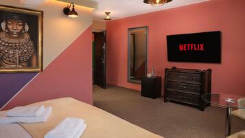 Habitación de hotel con una cama, una mesita de vidrio, un espejo, un televisor que muestra 'NETFLIX' y una obra de arte enmarcada en la pared.