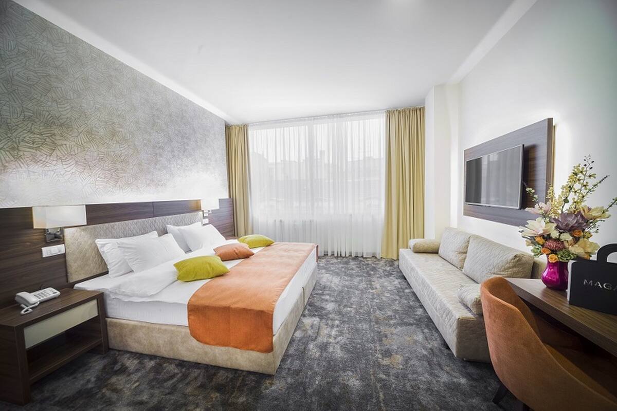 A hotel room with a large bed, a sofa, a desk with a computer, a TV, and a window with curtains.