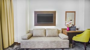 A hotel room with a beige sofa, a flat-screen TV mounted on the wall, a desk with a magazine, a vase with flowers, and a yellow chair. - Hotel Marshal (Belgrade)