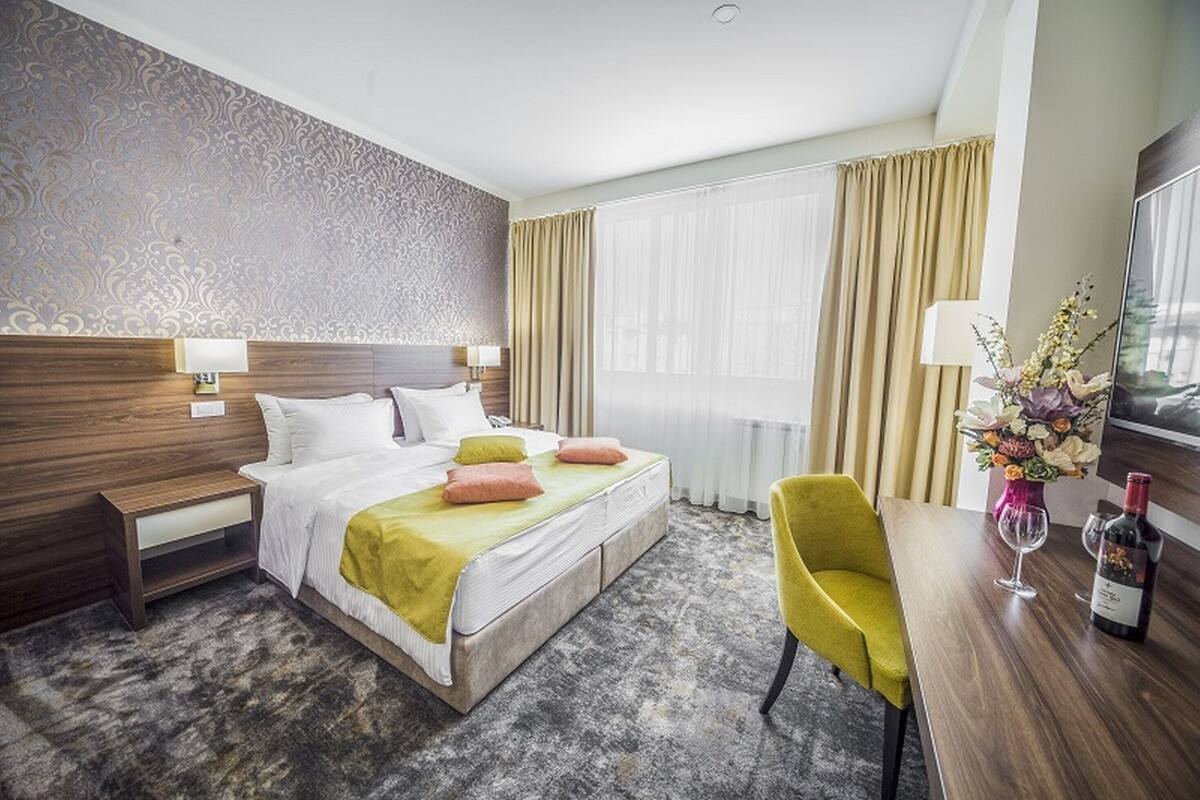 A hotel room with a bed, bedside tables, a desk, a chair, a vase with flowers, and a wine bottle.