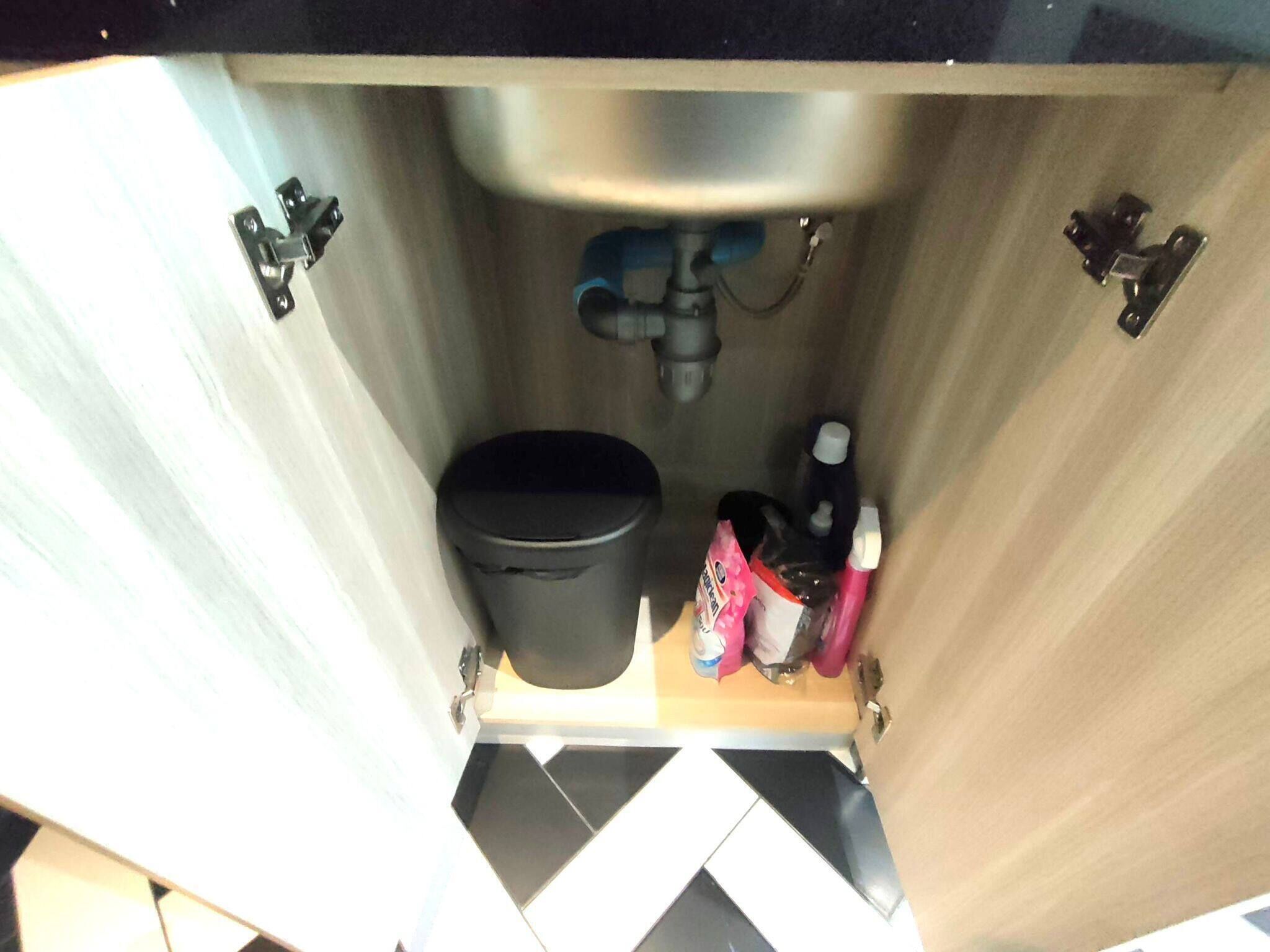 A kitchen sink with a black trash bin and a small shelf holding a bottle and a container.