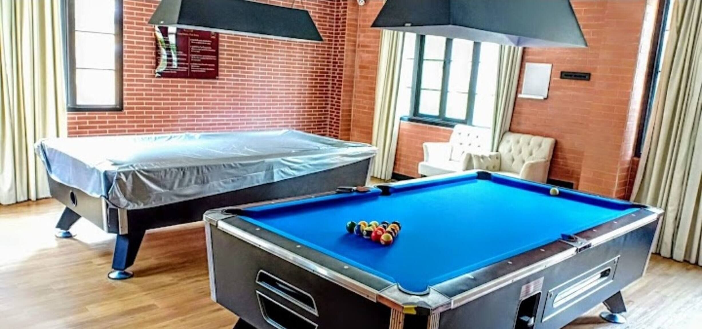 A pool table with balls, a covered sofa, and a brick wall with a window and curtains.