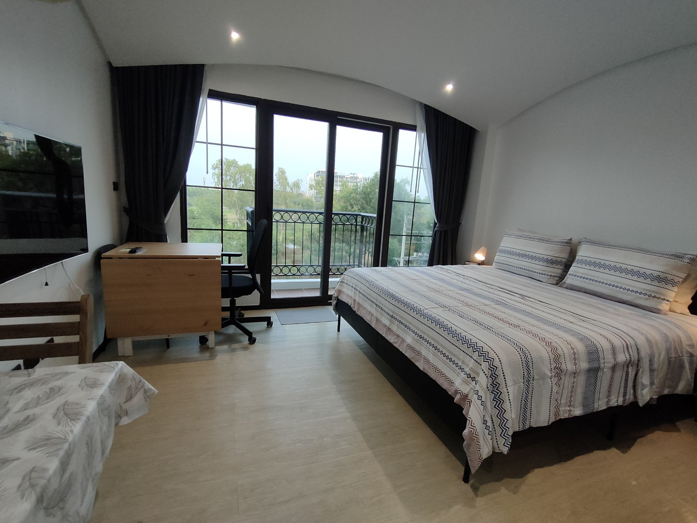 A bedroom with a bed, a desk, and a chair. There is a balcony with a view of greenery.