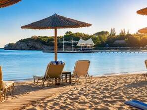 Une plage avec des chaises longues et un parasol en chaume, une jetée en bois et une côte rocheuse.