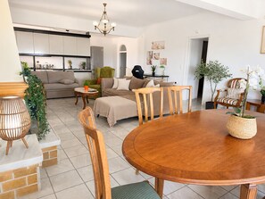 A modern living room with a dining table, sofas, and chairs. There is a kitchen area with cabinets and a refrigerator. - Eagles Nest Villa Your Grecian Paradise (Kropia)