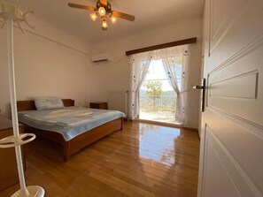 Un dormitorio con piso de madera, una cama con cubrecama azul, un ventilador de techo y vistas a través de una ventana con cortinas.