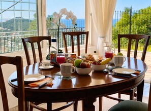 A dining table set with breakfast items, including fruits, pastries, and beverages, in a room with a view of the outdoors. - Eagles Nest Villa Your Grecian Paradise (Kropia)