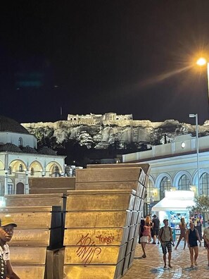 Vista nocturna de una ciudad histórica con una imponente fortaleza en la cima de una colina, una plaza pública con una gran escultura de metal y un edificio con arcos.