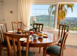 A dining area with a table set for breakfast, chairs, and a view of a mountainous landscape through a window. - Eagles Nest Villa Your Grecian Paradise (Kropia)